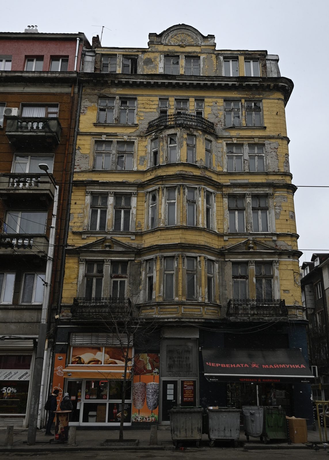 A crumbling yellow townhouse facade with broken windows above street shops