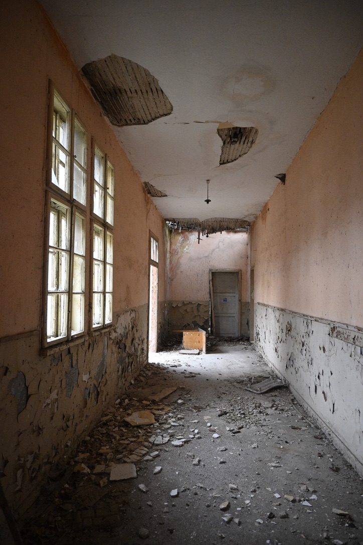 A peeling corridor with tall windows and a collapsed ceiling